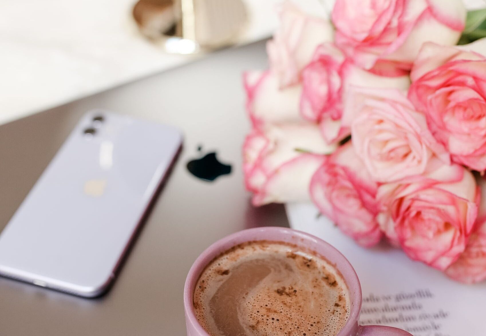 A delightful arrangement of a ceramic mug with cocoa, pink roses, a smartphone, and a laptop on a soft surface.